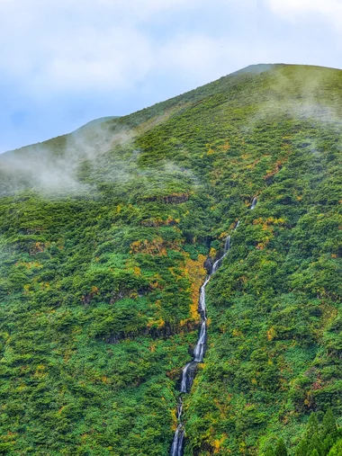 Waterfall cascading down a green hillside through mist and autumn foliage