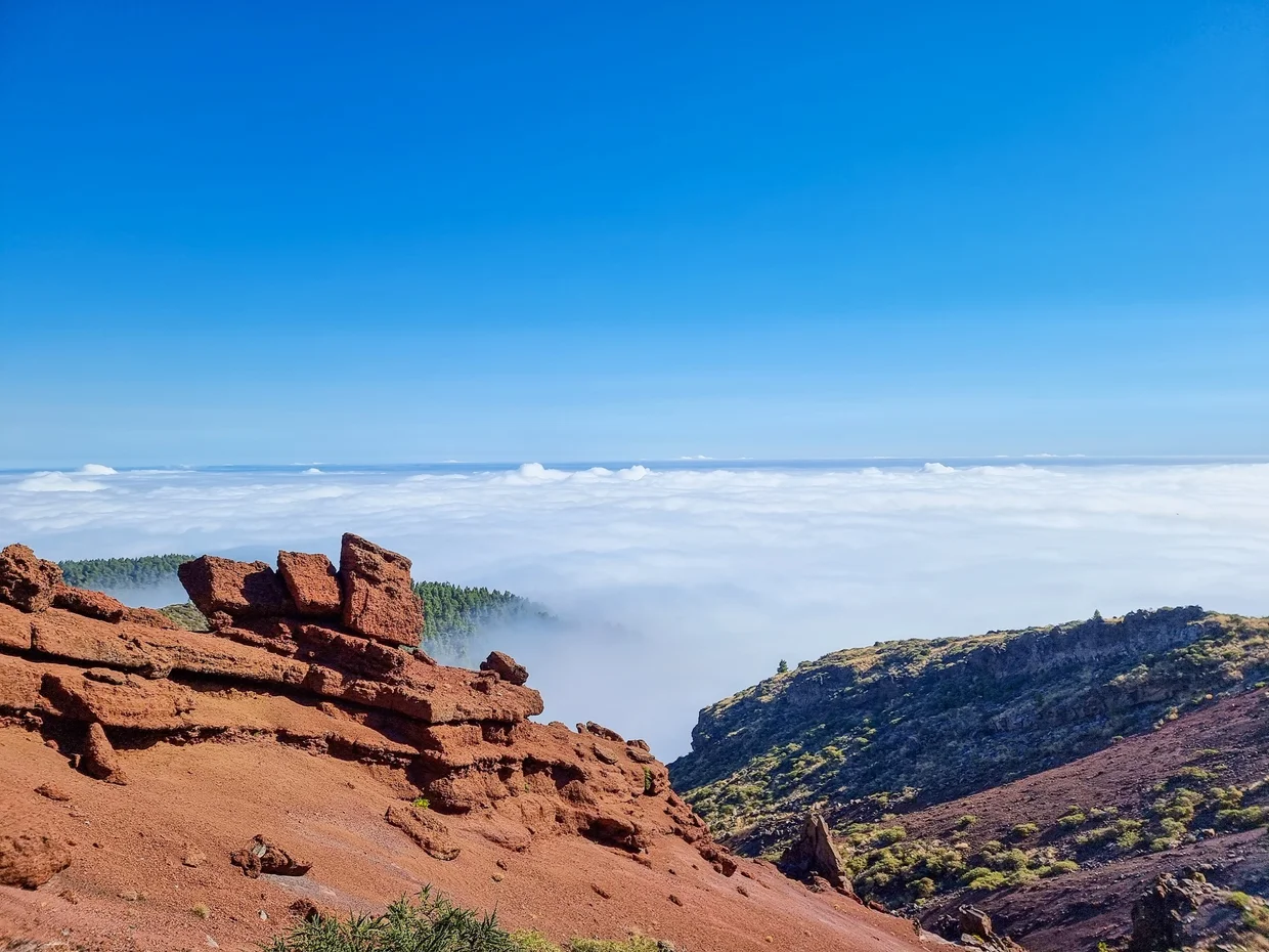 Red volcanic rock formations above a sea of clouds, clear blue sky